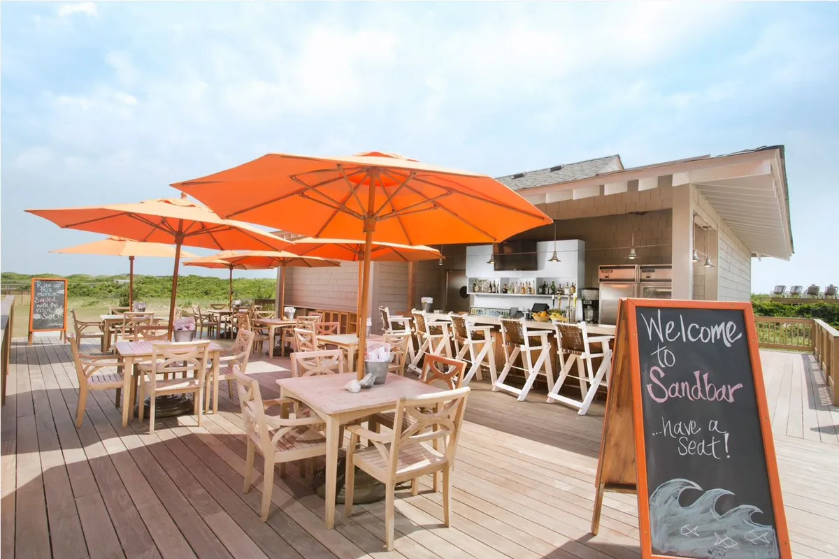 A cheerful outdoor dining area with bright orange umbrellas, wooden tables and chairs, a welcoming signboard, and an inviting bar area. The beach is visible in the background, reinforcing the casual, seaside vibe.