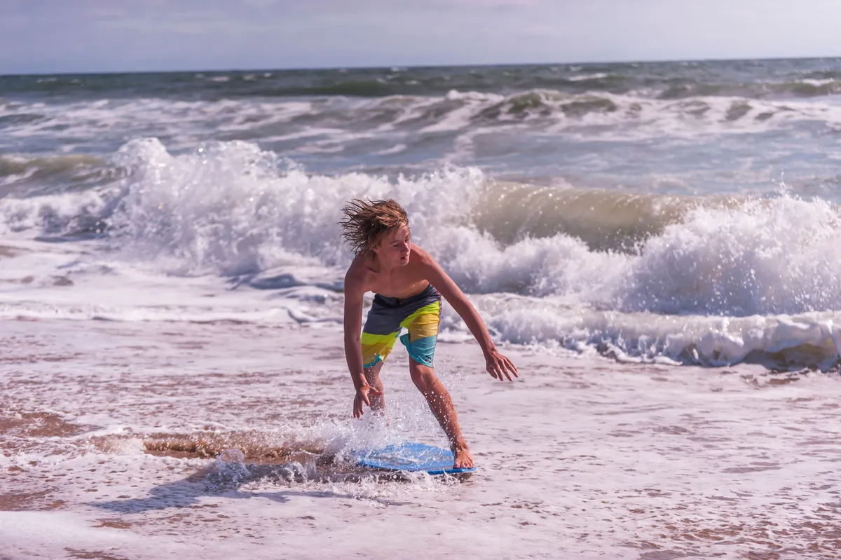 A young boy rides a boogie board on the waves, with the sandy beach and ocean stretching out behind him. The scene captures playful energy, with sandcastles in the foreground and splashes of water around the boy, evoking classic family beach fun.