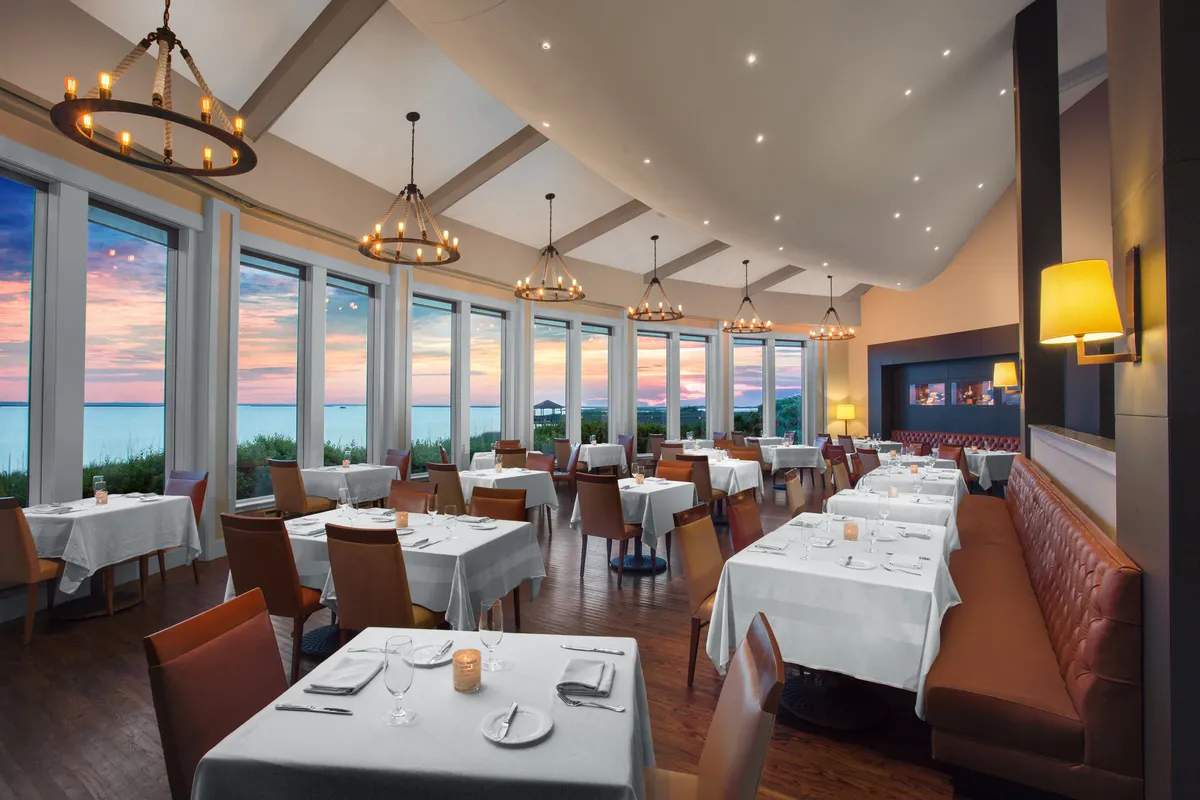 An elegant, softly lit dining space with large windows framing a vibrant sunset. Tables are set beautifully for an intimate meal, chandeliers provide a warm glow, and the overall mood is refined and romantic.