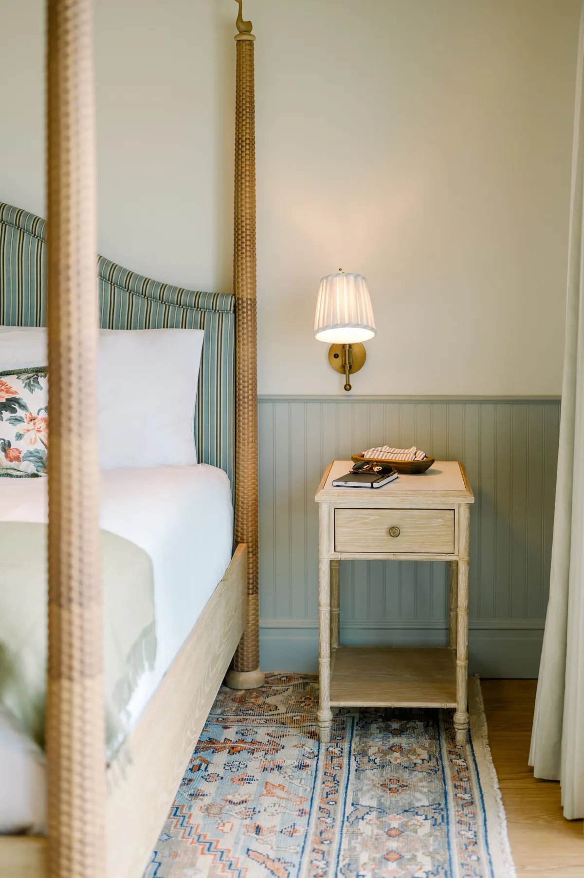 A serene interior space featuring a four-poster king bed adorned with decorative pillows and soft bedding, a nightstand with a lamp, and a wall sconce providing warm light. The scene evokes a sense of comfort and relaxation, inviting guests to unwind and rest.