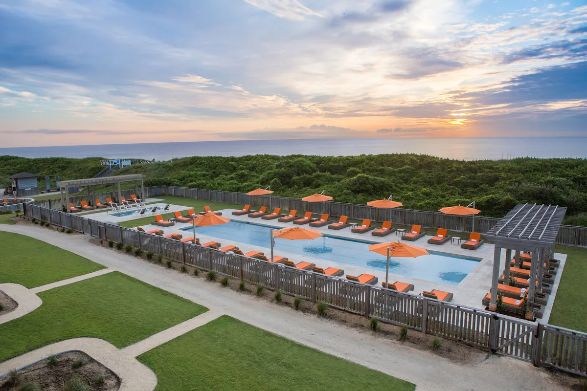 An outdoor pool scene at sunset, featuring a sparkling swimming pool surrounded by lounge chairs and vibrant orange umbrellas. The setting is tranquil yet inviting, ideal for swimming, sunbathing, and gathering for evening events.