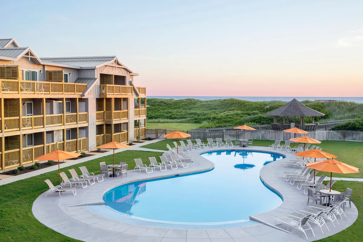 Expansive pool area surrounded by orange umbrellas and lounge chairs, with glimpses of buildings and a gazebo in the background. The tranquil atmosphere and inviting poolside setting showcase the view likely enjoyed from the balconies.