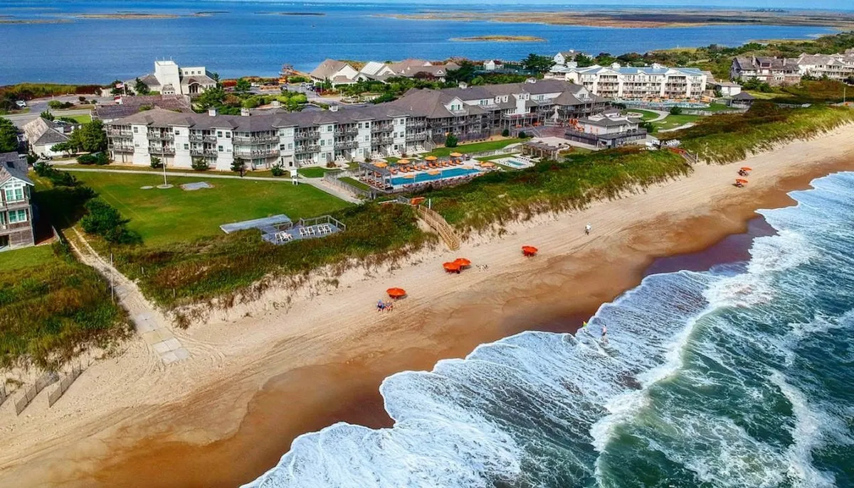 A sweeping view of the resort's lush green lawn, hotel pool, and orange beach umbrellas set against the backdrop of the ocean. The vibrant outdoor scene showcases places for swimming, sunbathing, and relaxing, connecting the resort directly to the beach.