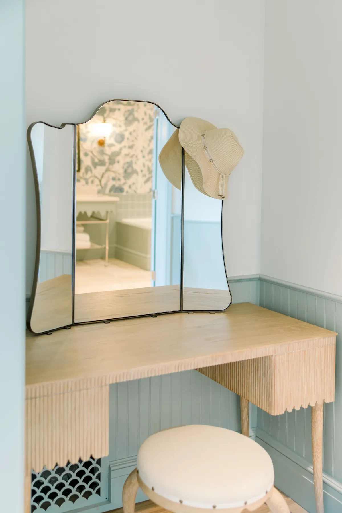 A tranquil corner with a wooden vanity table, stool, and a hat hanging on the mirror, suggesting a calm moment of personal care. The scene is intimate, highlighting thoughtful design and self-care elements.
