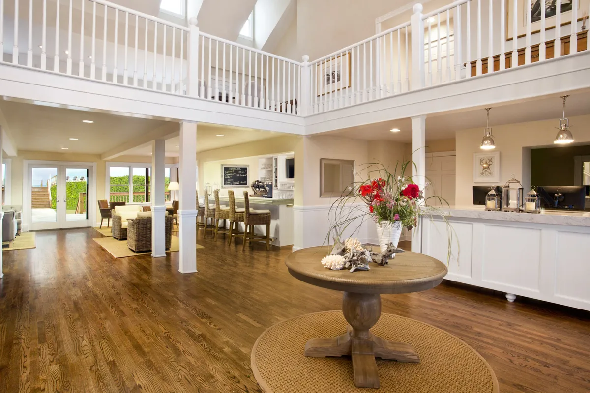 A spacious, sunlit room featuring a central round table adorned with a fresh floral arrangement, seamlessly connecting to an open kitchen with large windows providing views of the outdoors. The setting suggests a space designed for relaxed social gatherings, dining, and enjoying natural light.