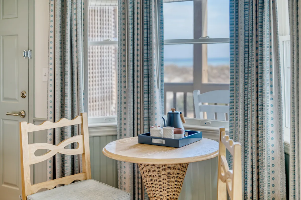 A cozy, sunlit nook featuring a small round table with a teapot and cups, a woven chair, and flowing curtains. The scene is set near large windows, providing a sense of seamless indoor-outdoor flow and inviting in natural light, perfect for enjoying a peaceful morning coffee or reading a book.