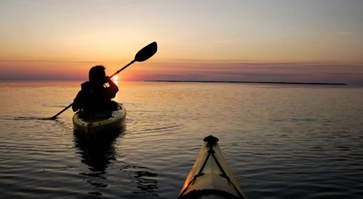 A kayaker glides across calm water at sunset, the sky painted in warm hues. The peaceful scene captures the spirit of outdoor adventure, with the kayaker surrounded by natural beauty and tranquility.