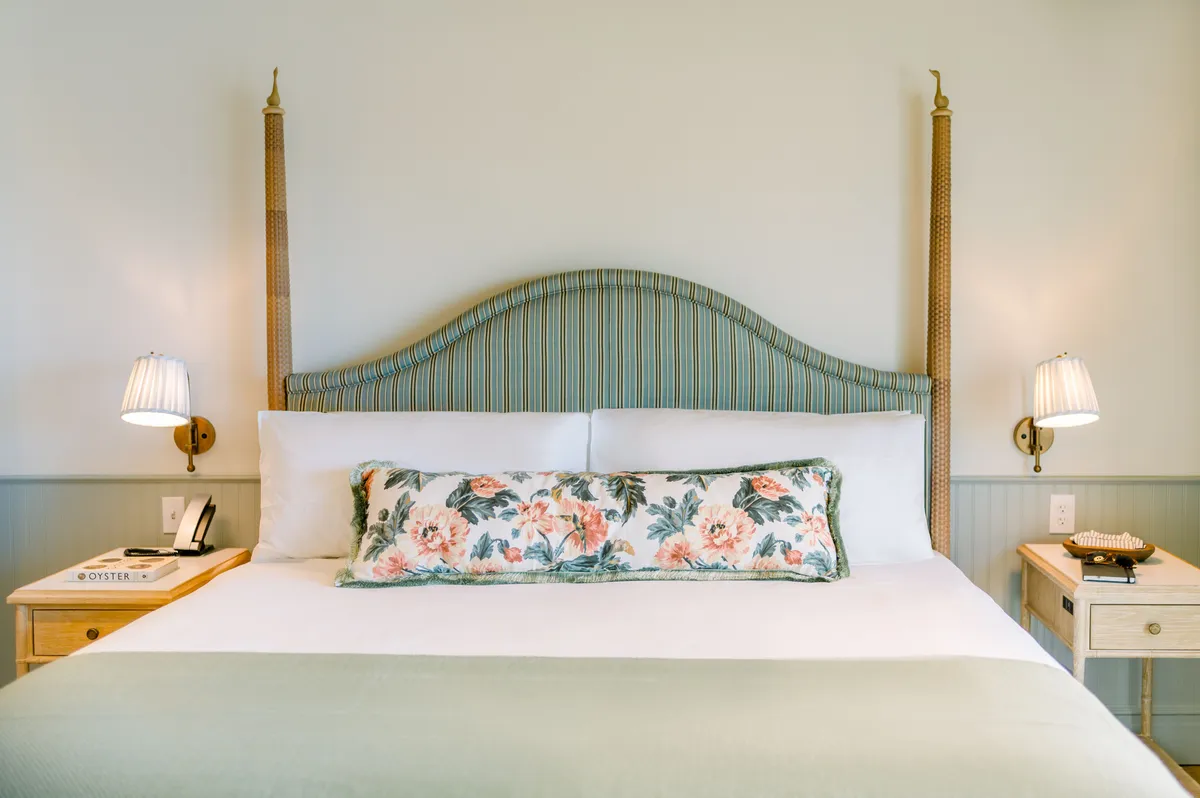 A tranquil bedroom corner with a comfortable bed accented by a floral pillow, a table lamp, and a nightstand. Natural light filters in, creating a peaceful, inviting space for rest or reading.