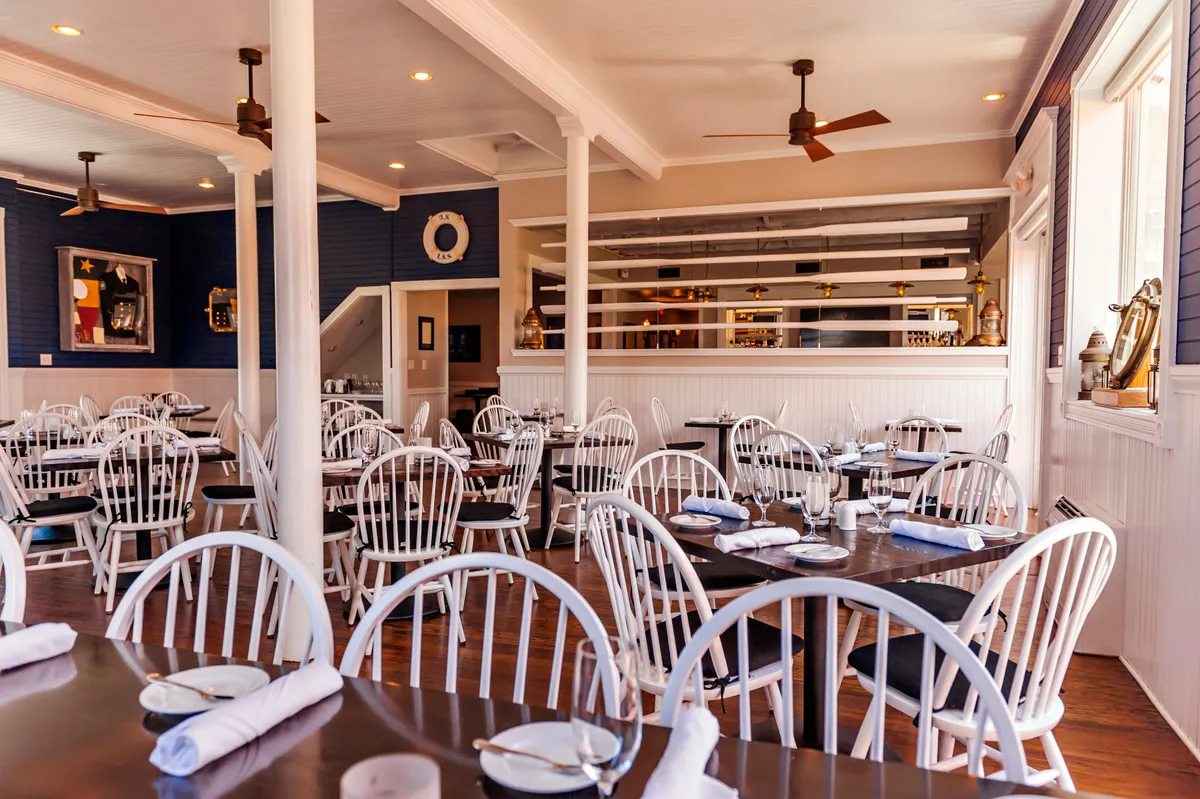 A cozy dining space with wooden tables, white chairs, a bar area, and stylish wall art. The ambiance is casual yet elegant, well-suited for meals with friends or family and celebrating special moments.