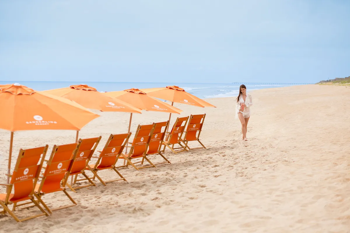This image captures a serene stretch of private beach with inviting orange beach chairs and umbrellas. A woman casually strolls along the shoreline, while the sky hints at sunset colors. The tranquil setting emphasizes relaxation and a romantic seaside experience.