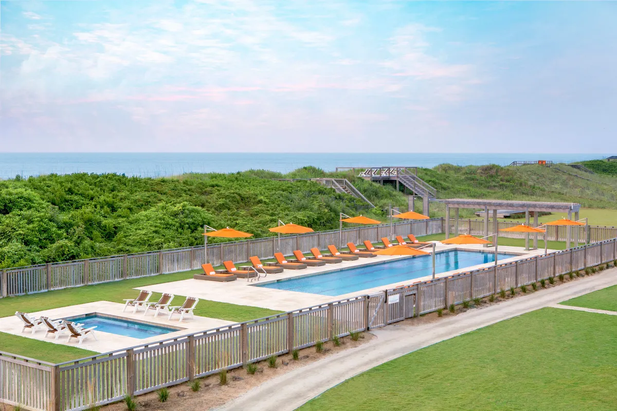 A bright, inviting outdoor pool area with orange lounge chairs and lush landscaping. Access stairs lead down to the beach, suggesting easy transitions between water, sun, and sand. The scene is vibrant yet serene, emphasizing the resort's commitment to relaxation and wellness.